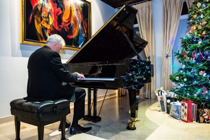 Steve Pietkewicz plays grand piano next to a colorful abstract painting. and a trimmed Christmas tree with packages underneath.