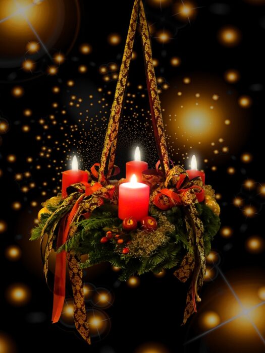 Advent wreath with four lighted red candles hangs suspended from a rope with greenery around it and sparkling star lights against the darkness.