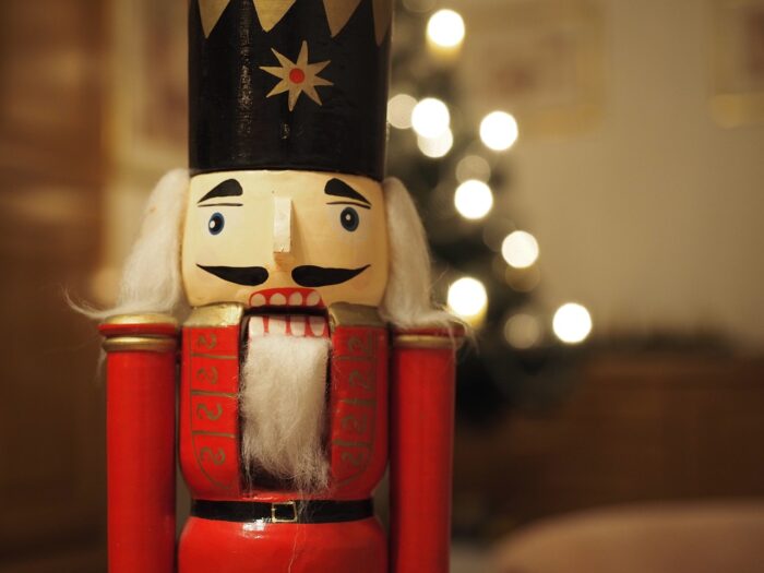 Wooden nutcracker in closeup with lighted Christmas tree blurred in the background behind him.