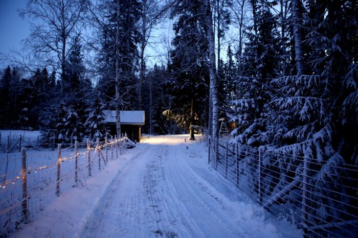 Small house at the end of a snowy road with lights on te fence
