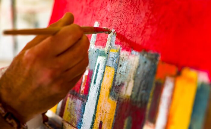 Closeup on an artist's hand with a paintbrush, as they add a red sky above a cityscape.