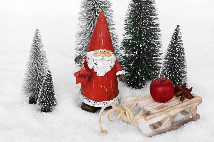 Santa resin ornament with white beard stnads near green miniature pine trees and a wooden sled with an apple on it.