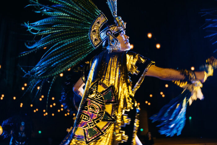 Sal Lopez, in Aztec-patterned gold robe and full Aztec headdress with blue feathers, onstage with gold lights in the background.