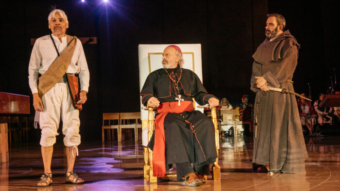 Scowling priest in full robes and sash sits on a chair between a monk and a man dressed in peasant clothes.