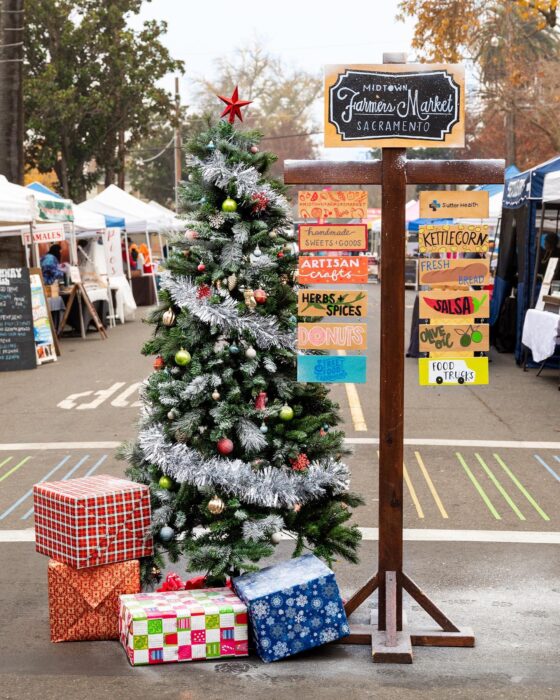 Farmers Market sign near Christmas tree trimmed with tinsel garlands and with packages under it, with white vendor tents in the background.