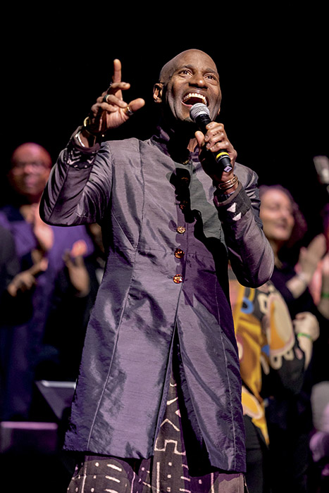 Vernon Bush, in a long jacket with black-and-gold trousers, sings into a mic with musicians in the background onstage.