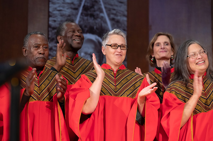 GLIDE Ensemble members, in red choir robes, smile and clap their hands in time to the music.