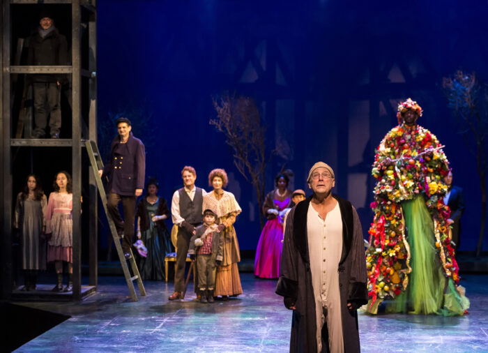 A Noise Within's cast of A Christmas Carol, with Scrooge front and center in his nightcap and nightgown, on a wintry stage.