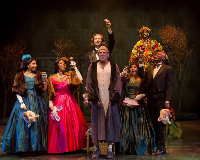 A bemused-looking Scrooge stands amid finely-dressed party guests cheerfully offering a toast as the Ghost of Christmas Present looks on.