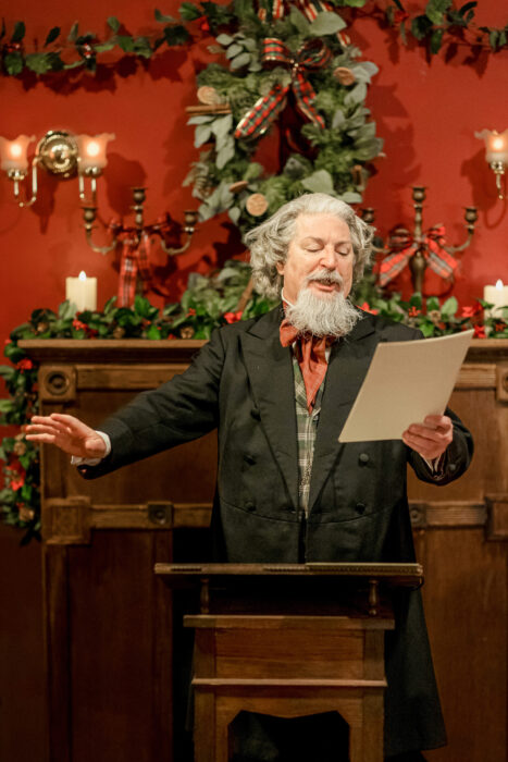 "Charles Dickens" stands at a podium, gesturing as he reads from his manuscript in front of a fireplace decke out with candles and greenery.