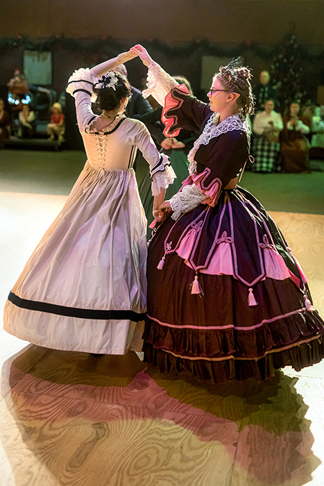 Two women in Victorian dress dance in a circle during Fezziwig's Dance Party.