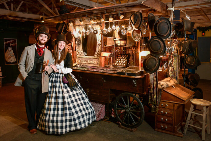 Couple in Victorian dress next to a lighted market cart showing handcrafted wares.