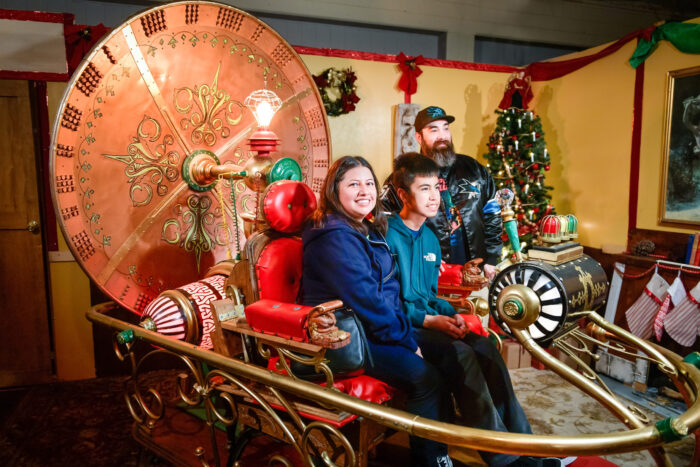 Couple ride in a "time machine" contraption with wheels and dials while inventor "Dr. Murillo" stands in the background near a Christmas tree trimmed with white candles.