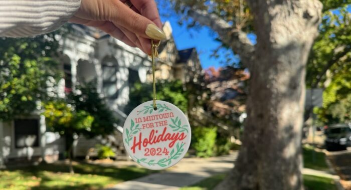 Woman's hand with painted nails holds a ceramic Christmas ornament with "In Midtown for the Holidays 2024" printed on it, with a white building in the background.)