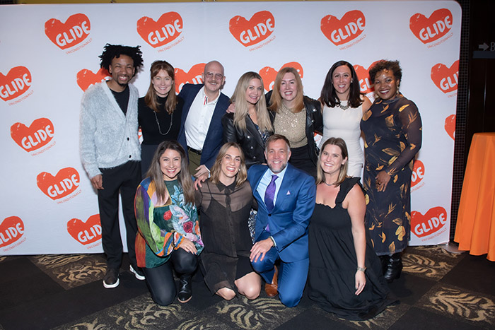 GLIDE staff pose in front of a white screen covered with red hearts with "GLIDE" in each, in white letters.