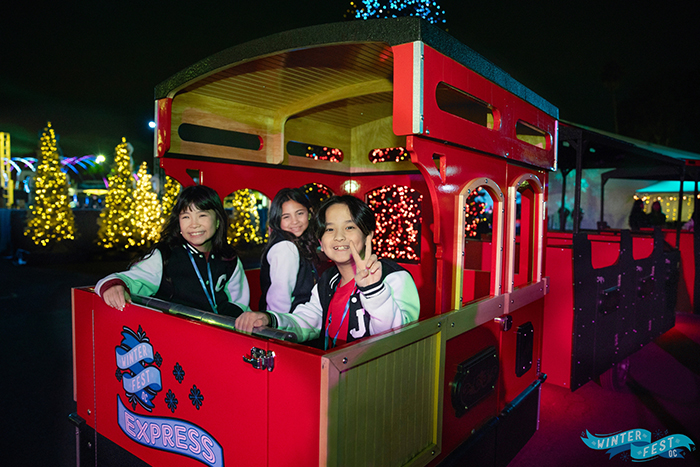 Laughing small girl flanked by two women in a red Christmas train with "Winter Fest OC" graphic in one corner of the screen.