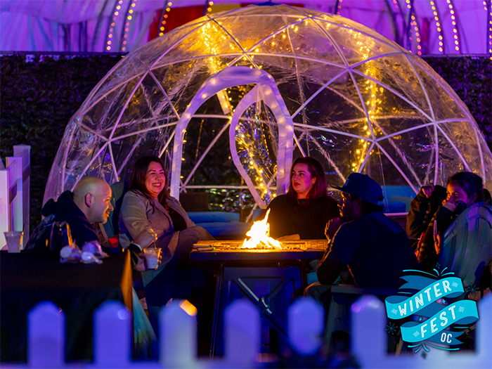 Adults gather around an outdoor fire it with gold lights in an archway behind them.