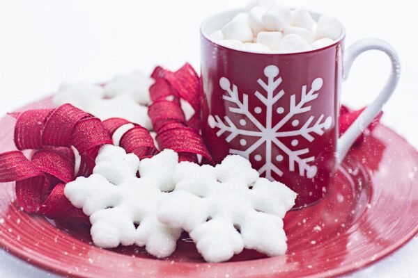 Decorated "snowflake' sugar cookies on a red tray next to a red mug of hot chocolate with a white snowflake on it and marshmallows up to the brim.