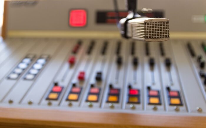 Red on-air light glows over a board with lighted busses and sliders up near a unidirectional mic.