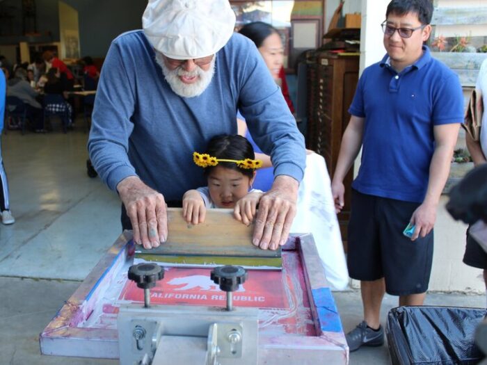 Bearded older man helps a small girl silkscreen a T-shirt.