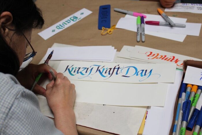 Young woman inks "Krazy Kraft Day" in blue calligraphic writing.