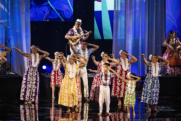 Daniel Ho plays his ukelele as he stands onstage, elevated above brightly-dressed hula dancers