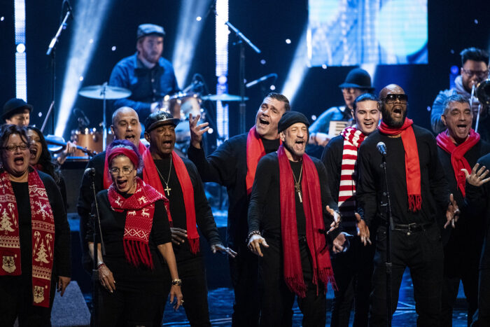 Urban Voices Project choir, wearing jackets and red or striped Christmas scarves, sings onstage in front of a drummer at his kit