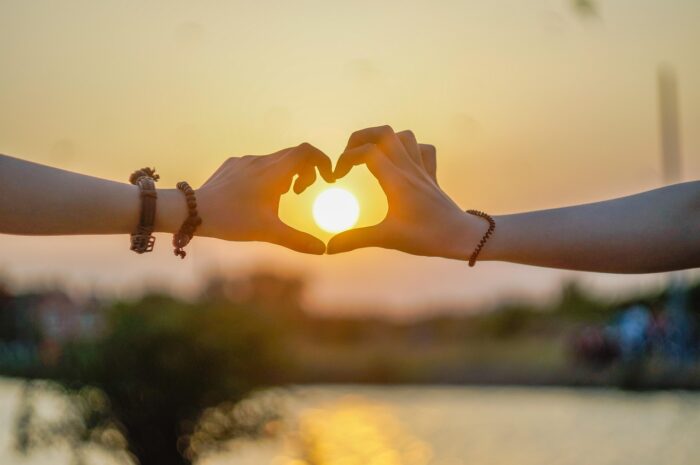 Two hands make a heart shape to frame a sun rising over a lake.