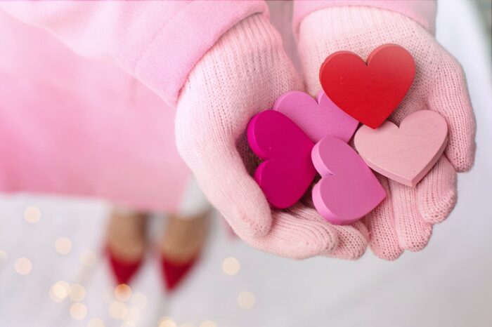 Woman's cupeed hands, in pink woolen gloves, offer five wooden cutout hears in red and pink. Pointed high-heeled shoes are visible in the background.