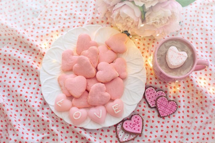 Pink iced heart-shaped cookies on a plate with the bottom four spelling out "L0O-B-E" and a cup of cocoa with a heart-shaped marshmallow.