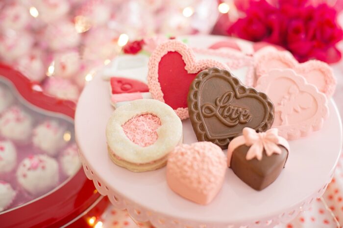 Chocolate-covered brown and pink heartshaped confections and white cookies with pink and red heart-shaped cutouts on a plate, surrounding a chocolate heart with "LOVE" printed on it, and red roses in the background.