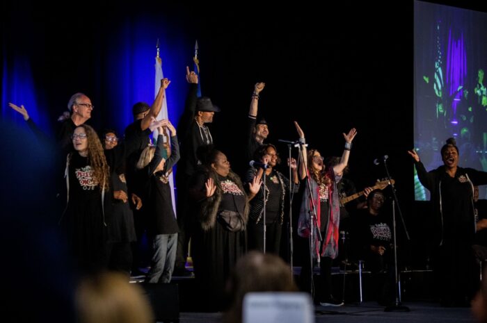Soloist raises her arms skyward as she sings onstage, with choir and American flag in the background