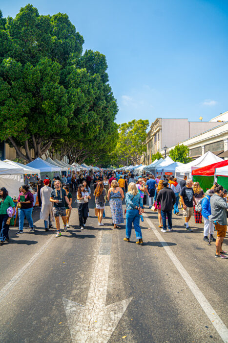 Shoppers browse a street of artisan booths under a blue sky with green trees and buildings in the background.