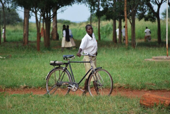 Young African man walks bicycle along a dirt path with grass along each side and trees and other students in the distance.