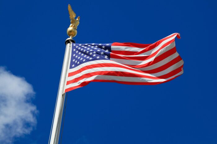 American flag with golden eagle atop its flagpole waves against a blue sky with one cotton-puff cloud.