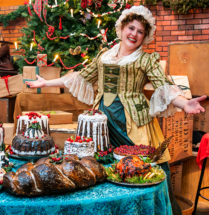 Mrs. Fezziwig smiles near a table laden with Christmas cakes and pudding.