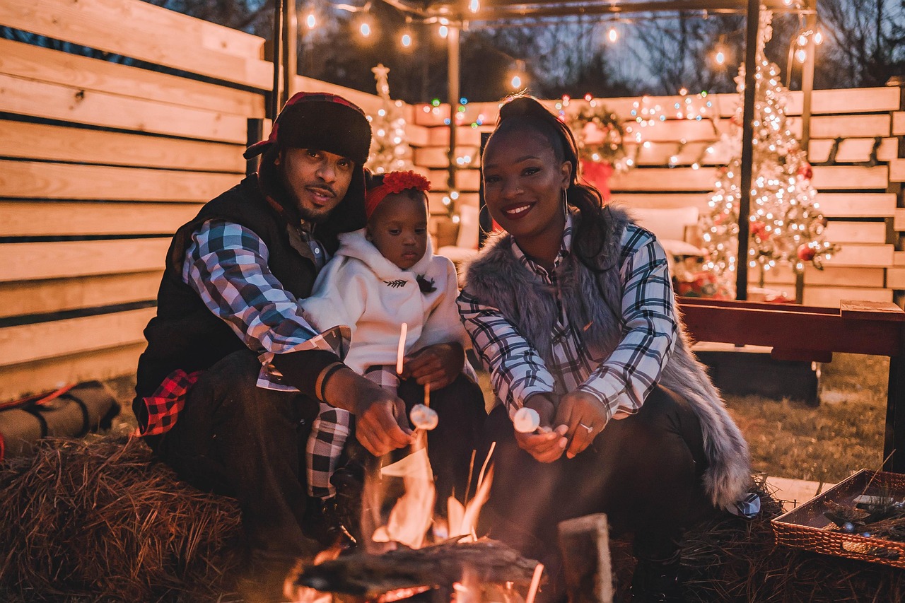 African-American husband and wife, with their baby, sit in a wooden structure decorated with Christmas lights and red ribbons and toast s'mores over a fire pit.
