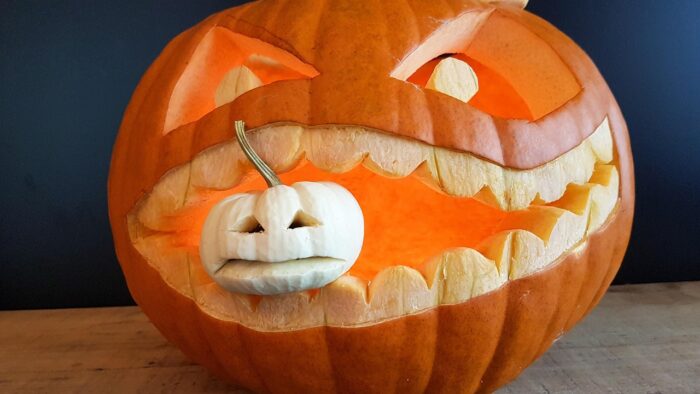 Large orange jack-o-lantern holds smaller white jack-o-lantern in its mouth.
