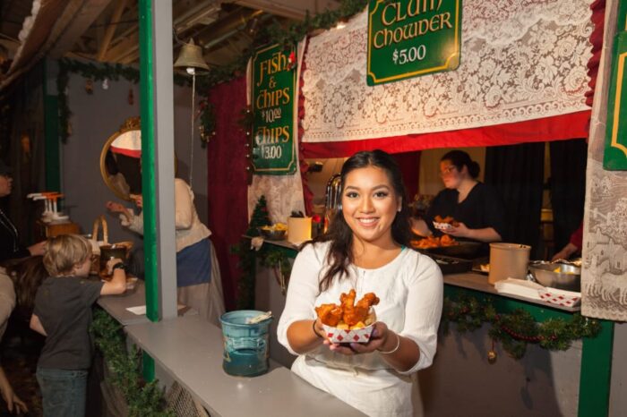 Smiling vendor at a booth adverting "Clam Chowder, $5" offers a cardboard container of battered fish and chips.