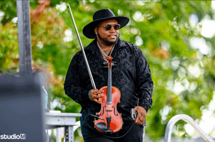 Dominique Hammons, in a black shirt, hat and shades, holds his classical violin and bow as he smiles against the background of a leafy tree and wicker outdoor chair.