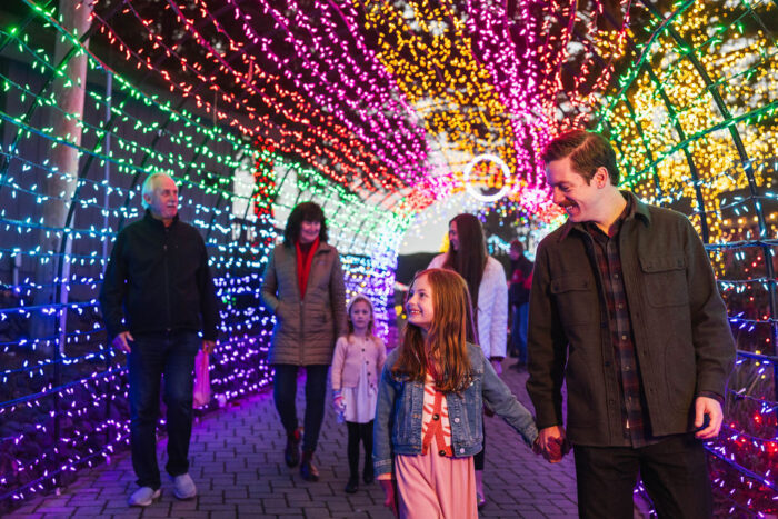 Father holds his small daughter's hand as other adults follow in the background through a tunnel of lights in rainbow colors.