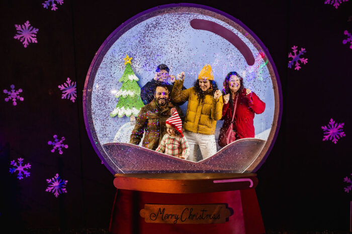 Family poses for a picture inside a "snowglobe" setting with green snow-covered trees against a Christmas-card background.