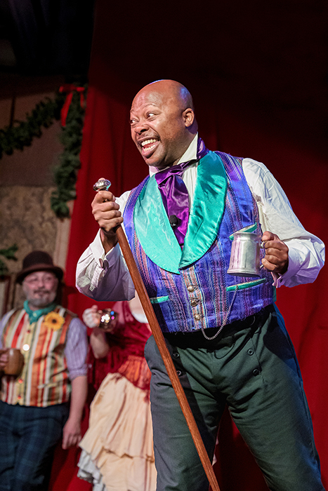 African-American singer in a purple waistcoat with blue lapes and a purple tie smiles as he grasps a standing mic.