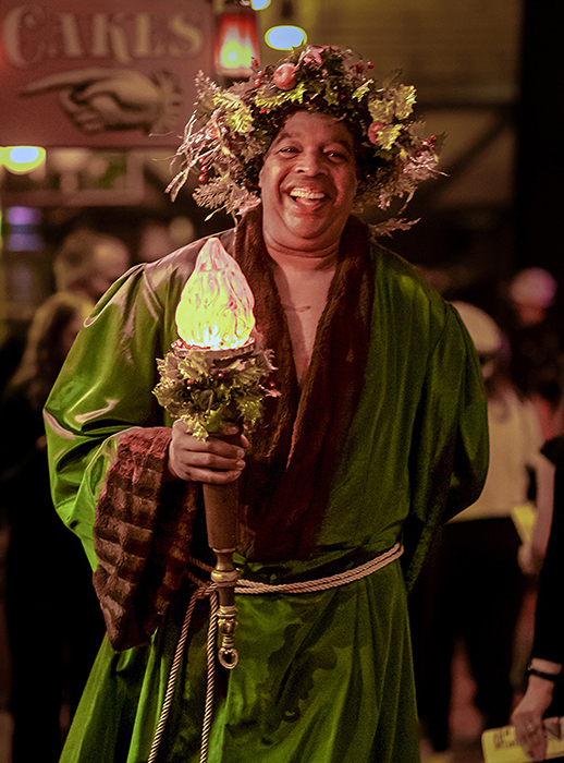 Ghost of Christmas Present, in a green velvet robe with a crown of gold holly leaves, smiles as he holds aloft a flaming electic "torch".