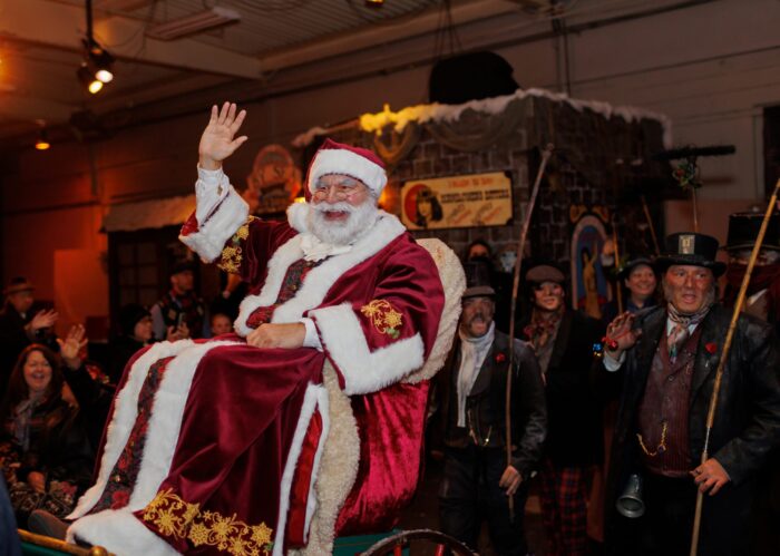 Father Christmas, in red robes trimmed with gold snowflakes and white fur, waves from his throne, surrounded by staffers in Dickensian clothing.