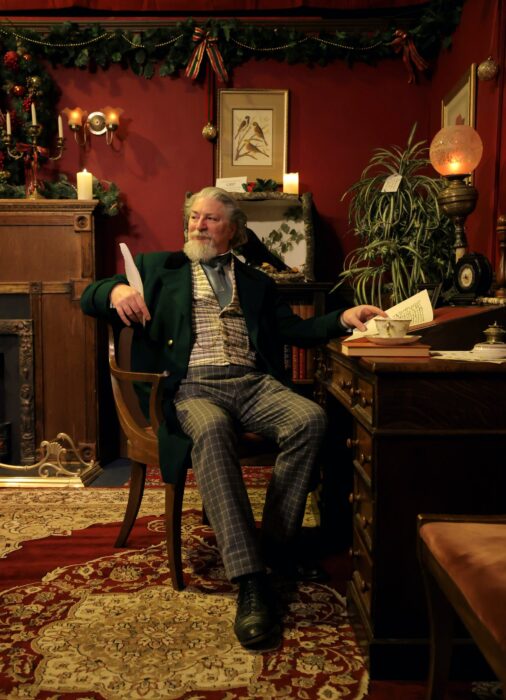 "Charles Dickens" sits at his desk in a Victorian study with an antique rug, a dark-wood fireplace and kerosene lamps.