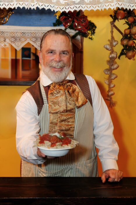 Bearded vendor with a gold brocade tie and a striped apron offers a cardboard bowl of pasta heaped with meatballs and tomato sauce.