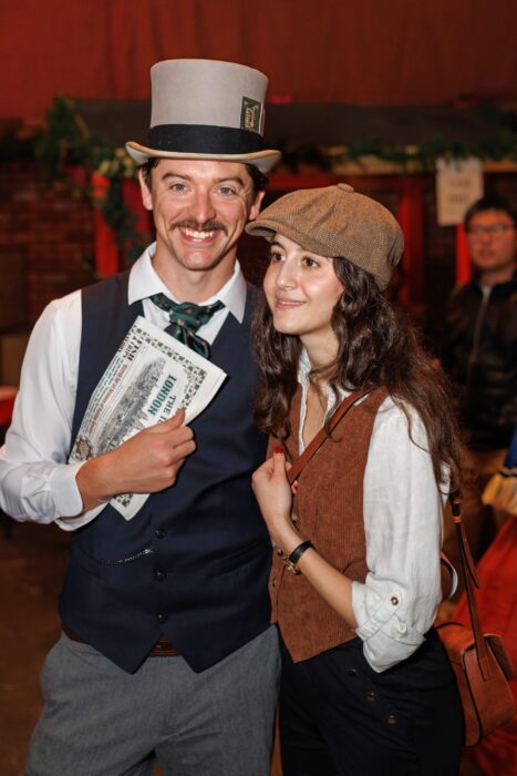 Young couple in Dickensian clothing: man in a top hat, holding a London newspaper, and woman in a newsboy's cap