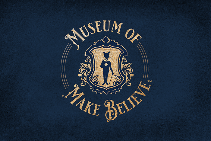 Museum of Make Believe logo with navy-blue background, cat in formal suit with cane in middle of a gold shield and "Museum of Make Believe" circling the shield in gold letters.