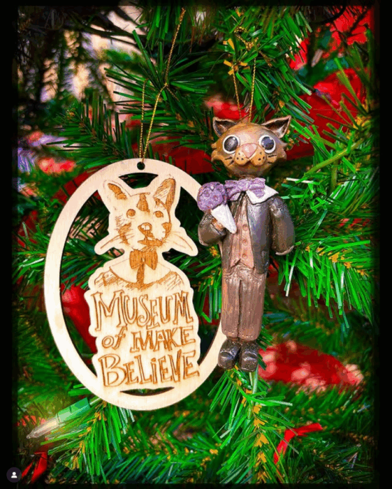 Wooden carved cat ornament with big eyes next to a wooden cat silhouette with "Museum of Make Believe" engraved into it.